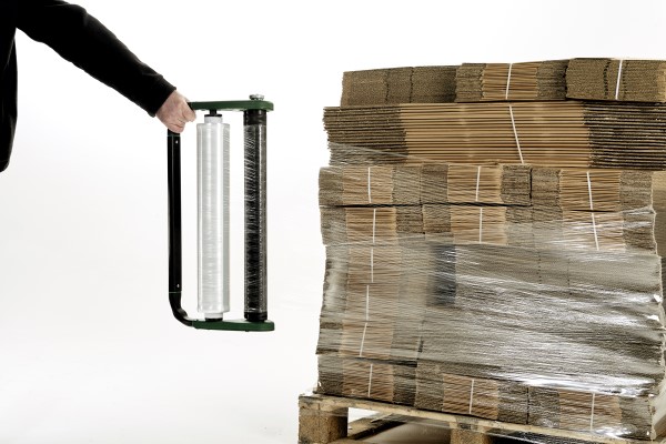 Person holding a manual stretch wrap dispenser next to a pallet of stacked and wrapped cardboard sheets.